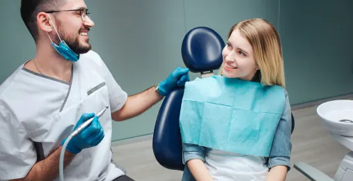 A smiling dentist with a handpiece talks with a young woman sitting in a blue dental chair