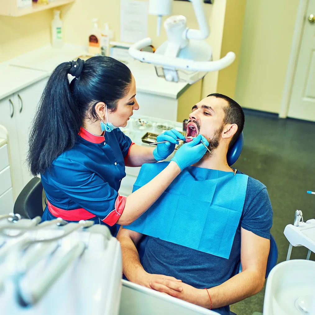 A dentist in a blue uniform examines a seated man's teeth