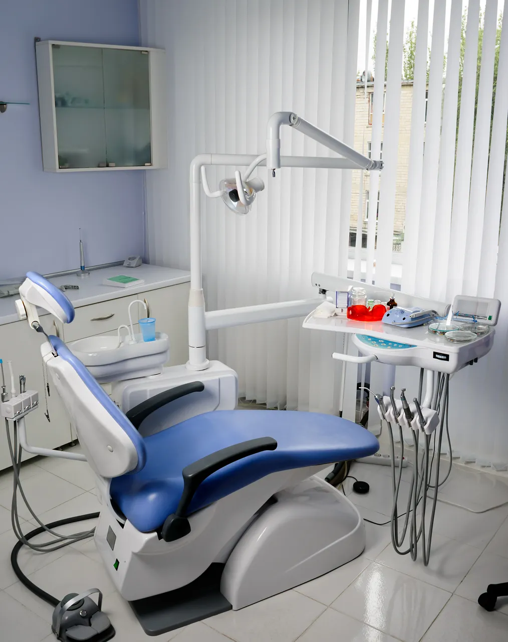 An empty treatment room with a blue dental chair, overhead lamp and instrument tray