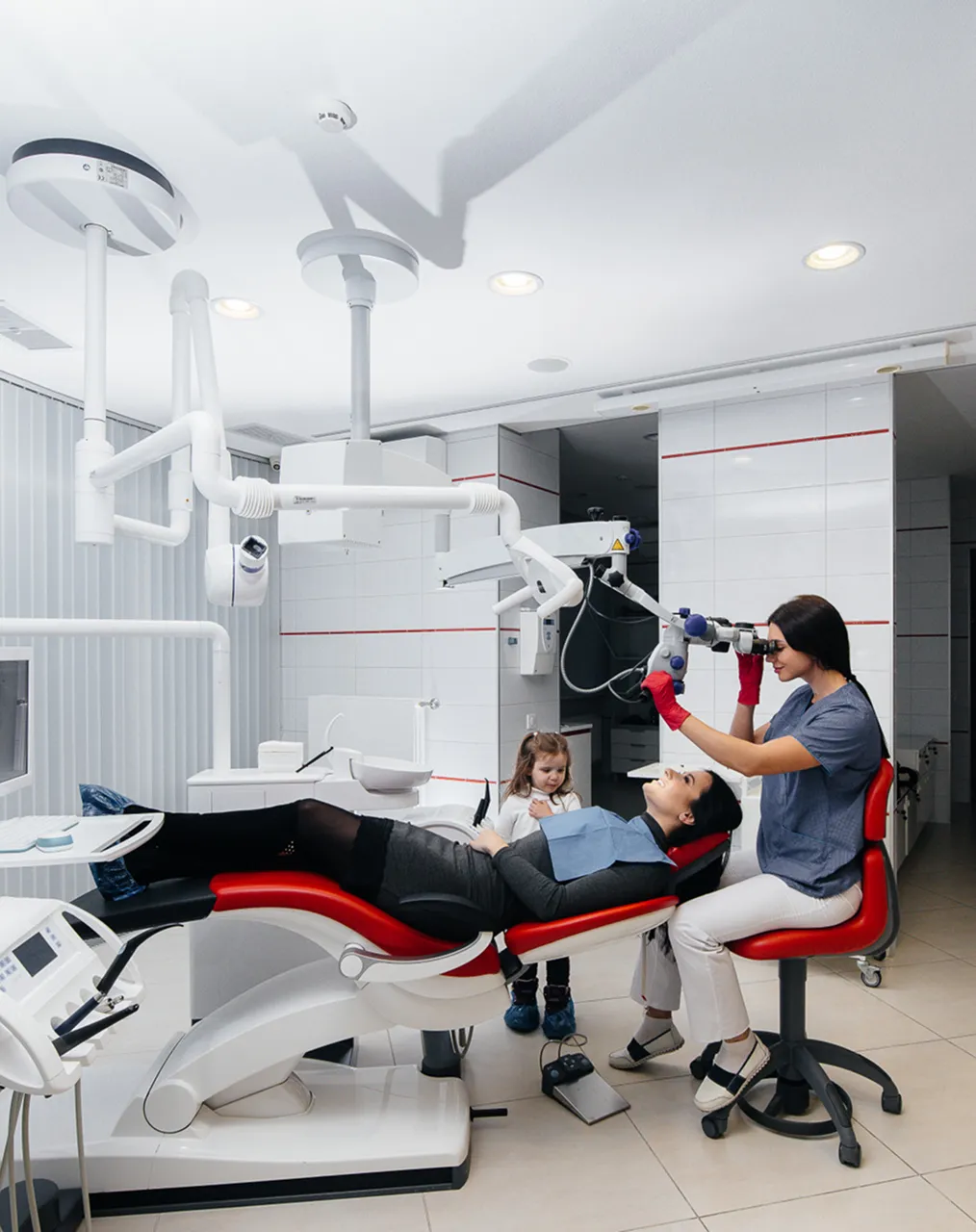 A wide view of a bright treatment room, a dentist at the microscope beside a reclined patient