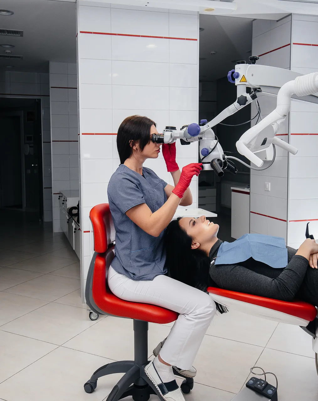 A dentist uses an operating microscope to treat a patient lying back in a red dental chair
