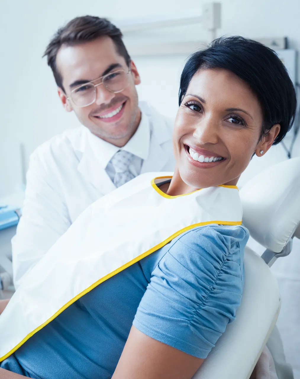A smiling patient in a dental bib sits in the chair, her dentist standing behind her