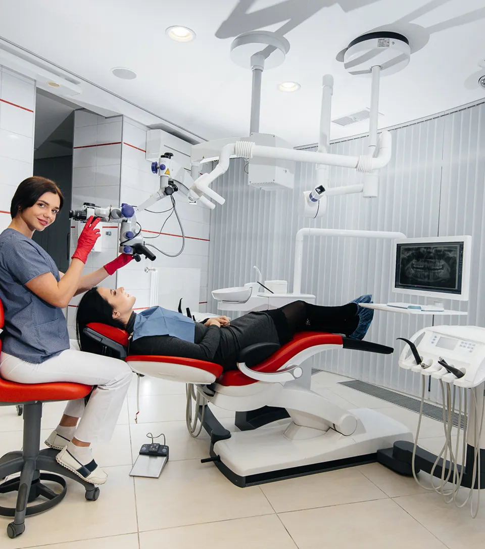 A dentist at an operating microscope treats a patient lying back in a red dental chair