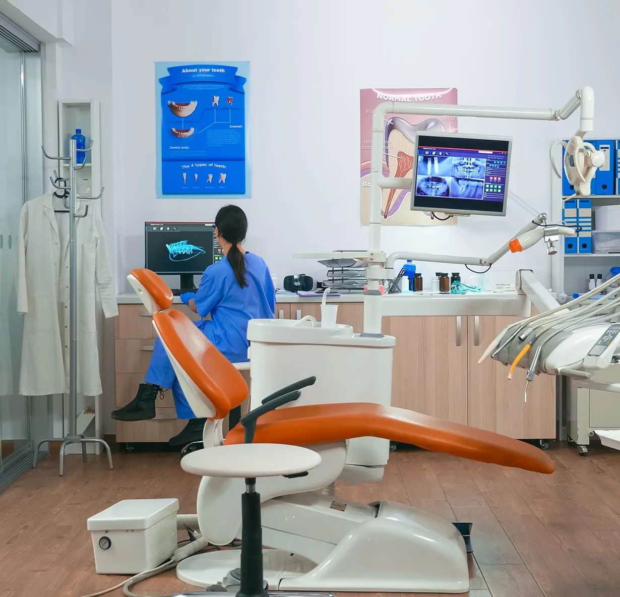 A treatment room with a dental chair, overhead lamp and screens, a staff member working at a desk behind it