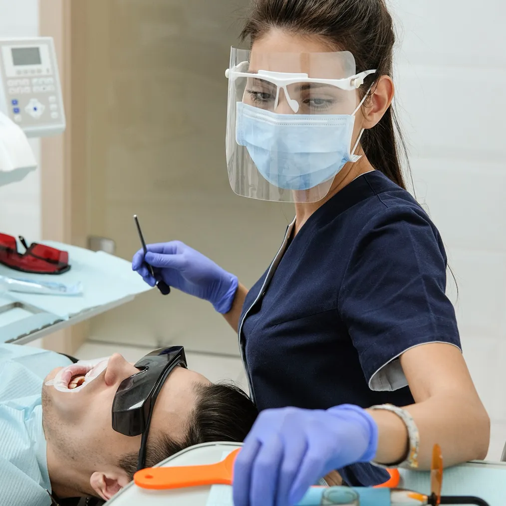 A dentist in a face shield, holding a dental mirror, treats a patient wearing protective glasses