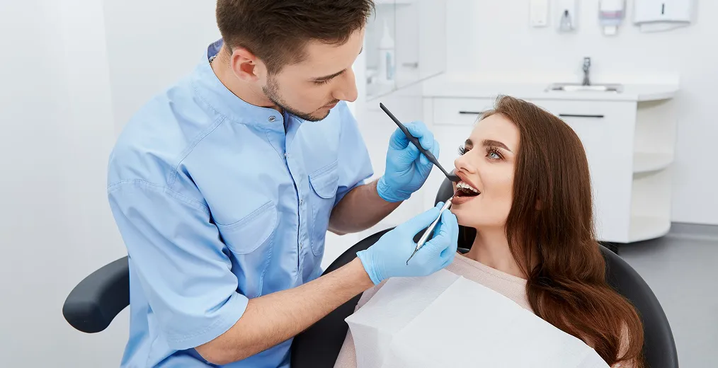 A dentist in a blue tunic examines a woman's open mouth with a dental mirror