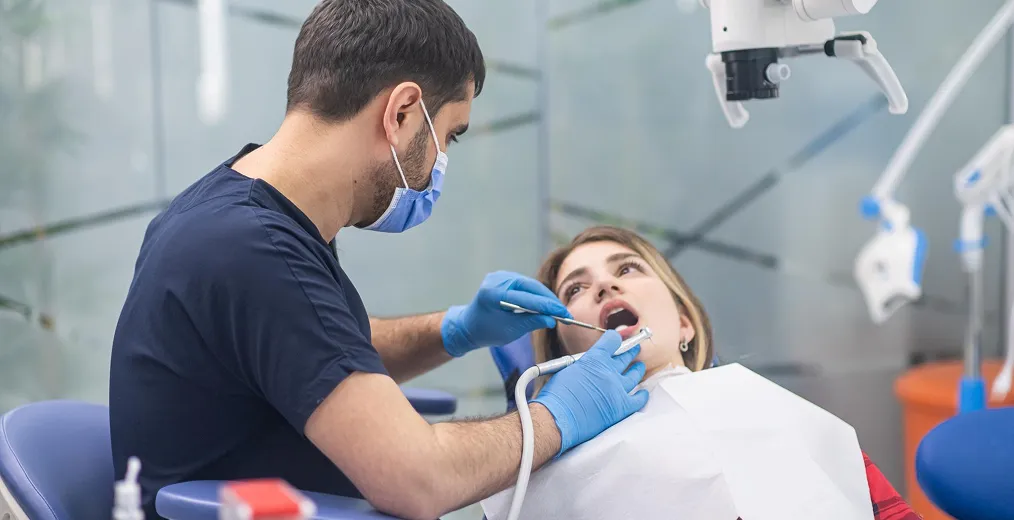 A dentist in a mask examines a young woman's teeth in the dental chair