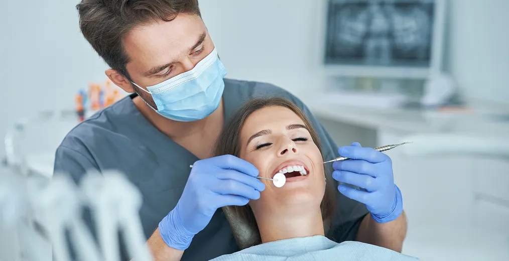 A dentist in a mask checks a smiling woman's teeth with a mirror and probe
