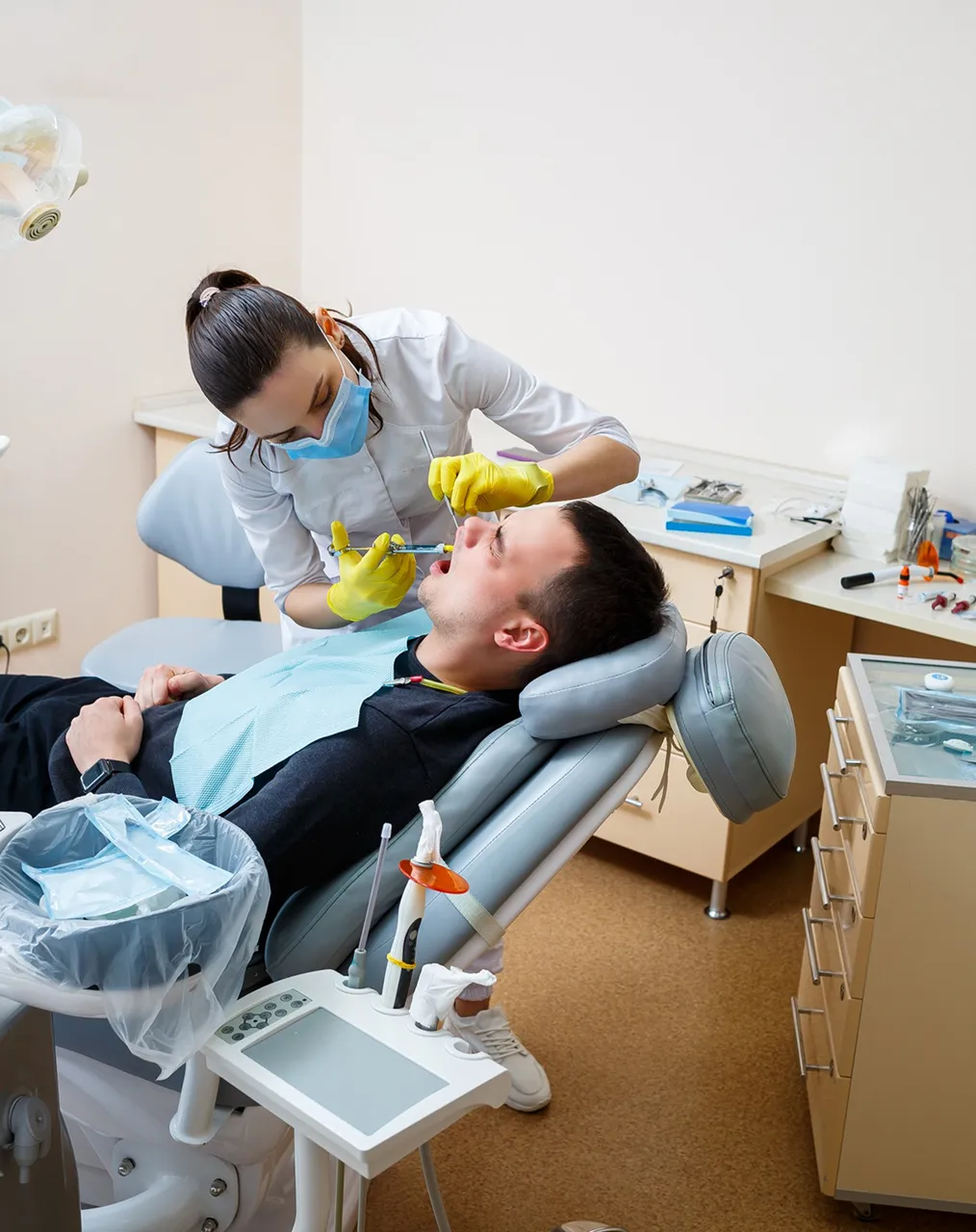 A dentist in a mask and yellow gloves works on a reclined patient while an assistant holds a suction tube