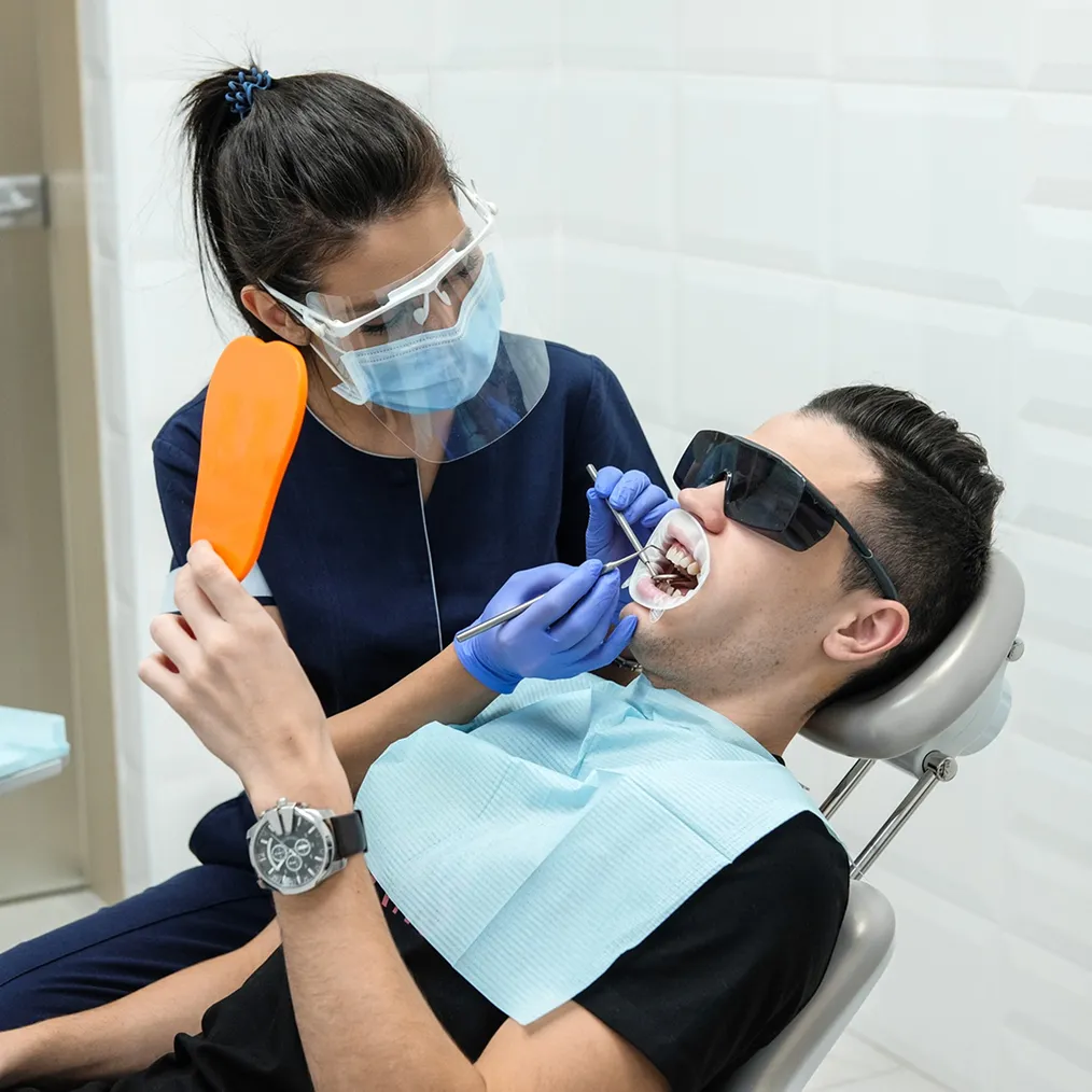 A dentist in a face shield and mask examines a young man wearing protective glasses, holding a curing light